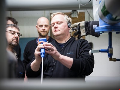 Tre tekniker i ett källarutrymme med rör i taket som studerar sin utrustning. 